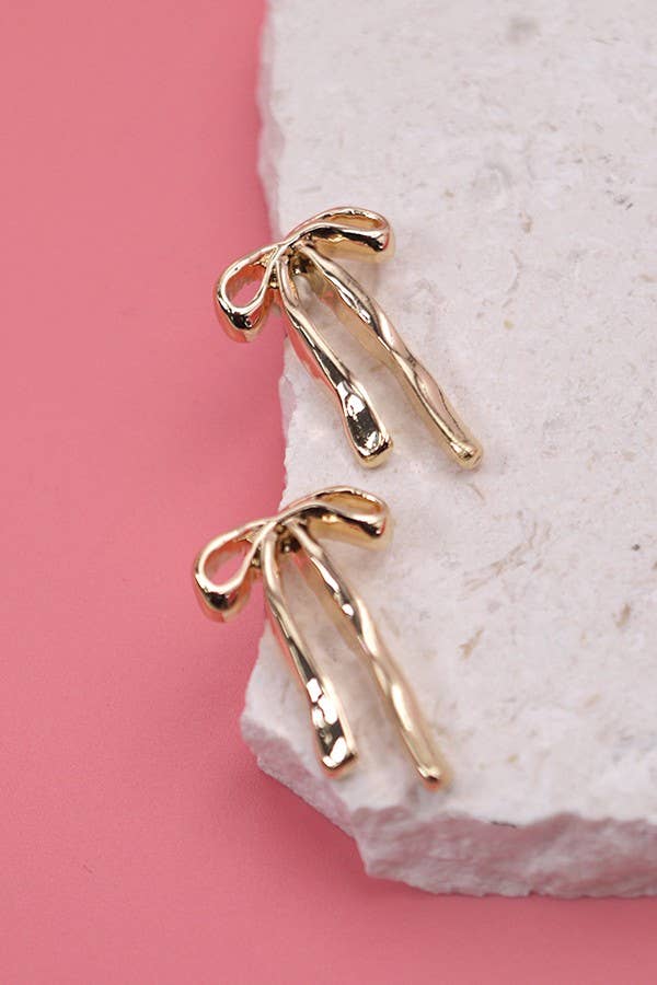Gold bow-shaped earrings on a stone surface with a pink background