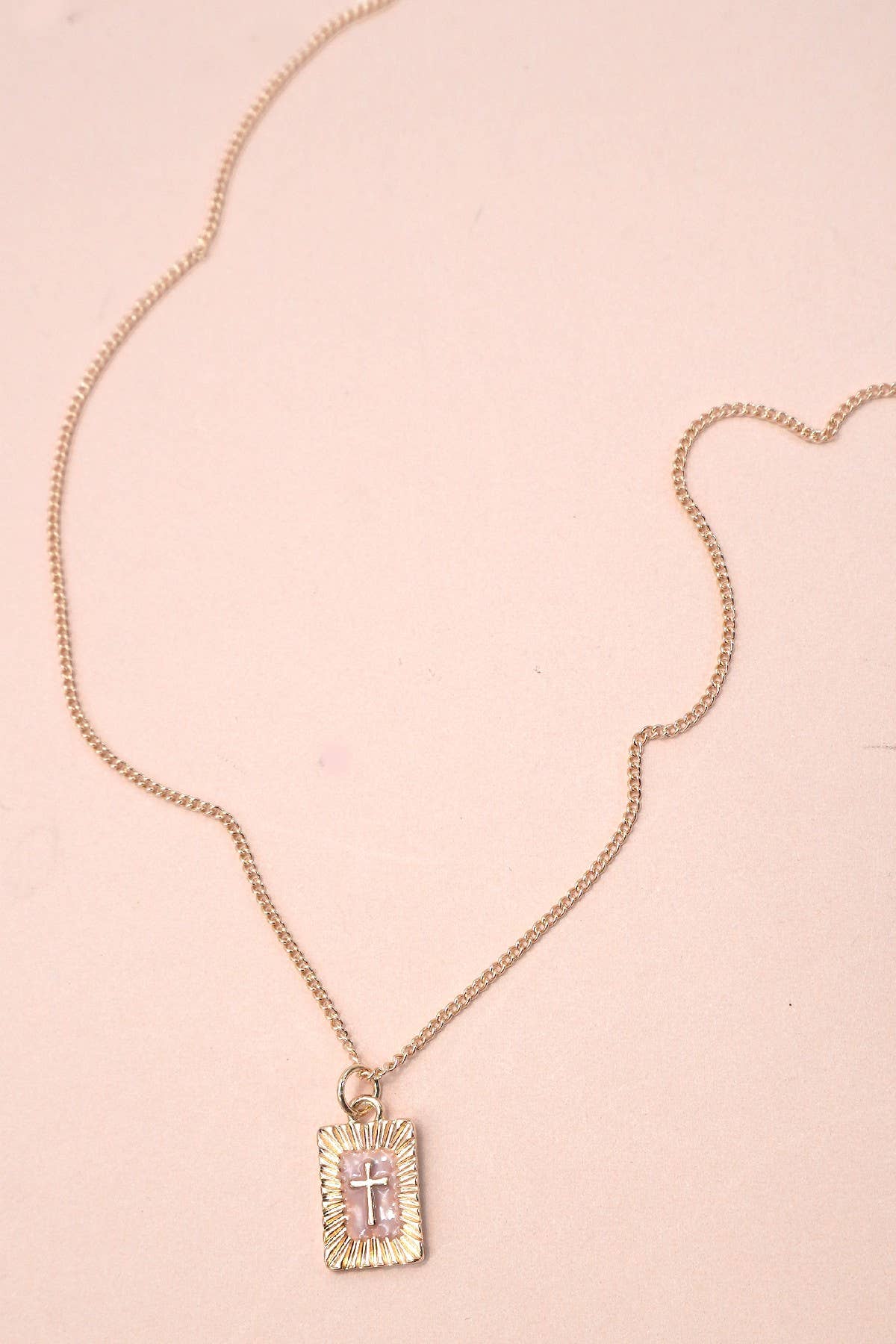 Gold necklace with a rectangular pink cross pendant on a pink background
