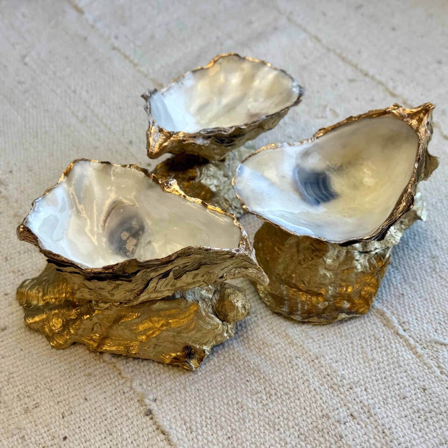 Three oyster shell ring dishes with gold interior linings on a textured beige surface