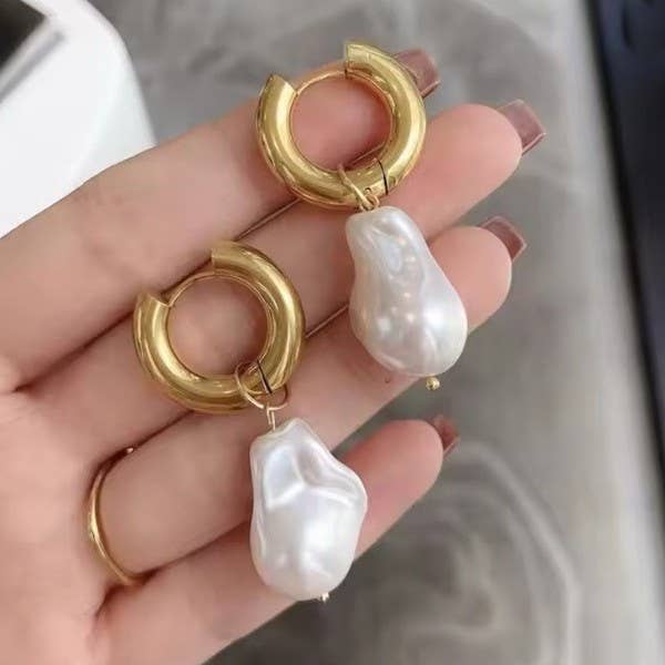 Gold hoop earrings with white pearls held in a hand against a neutral background