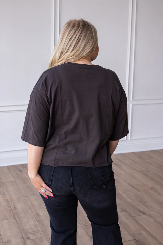 Person wearing a grey cropped shirt and dark pants in a room with wooden flooring and white walls.