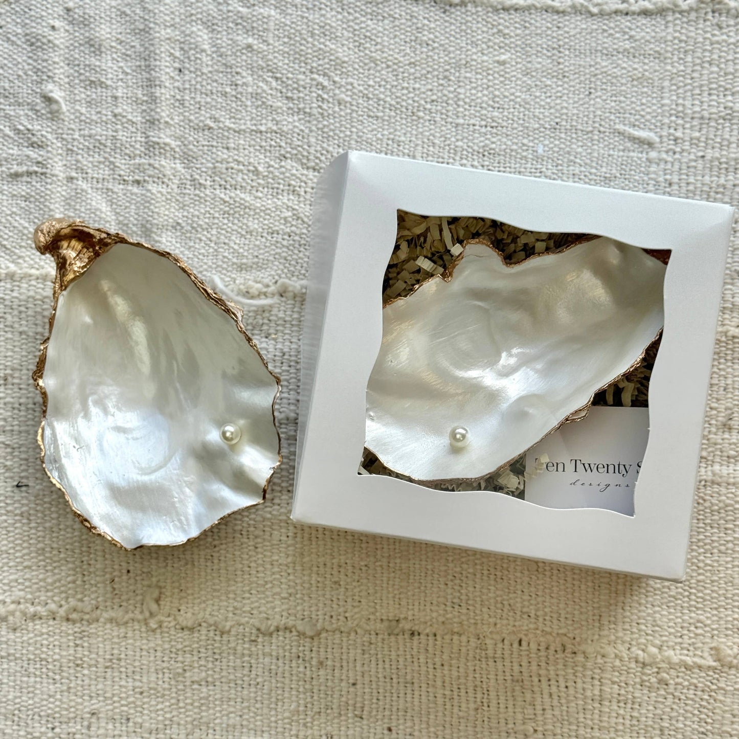 Pearl in an oyster shell jewelry dish with a branded box on a textured fabric background