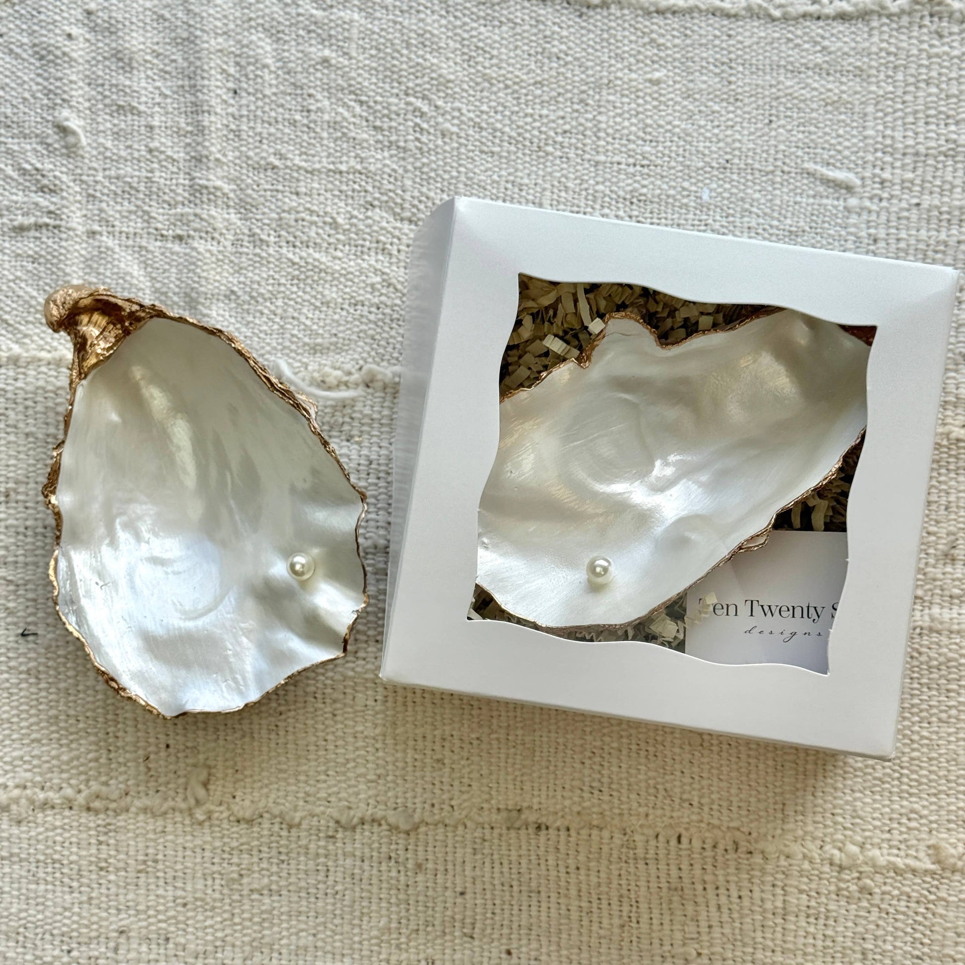 Pearl in an oyster shell jewelry dish with a branded box on a textured fabric background